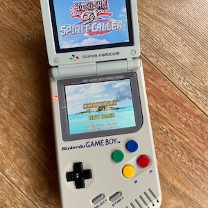 Puede incluir: Consola portátil Nintendo Game Boy Advance SP gris claro con dos pantallas. La pantalla superior muestra "Yu-Gi-Oh! GX Spirit Caller". La pantalla inferior muestra un menú del juego. Cuenta con botones de colores y una cruceta.