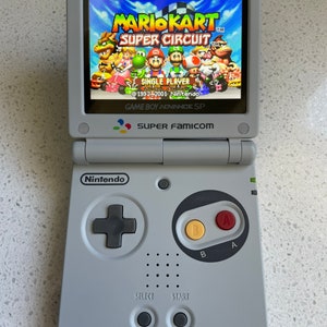 Puede incluir: Consola Game Boy Advance SP gris claro mostrando el juego Mario Kart: Super Circuit. La pantalla muestra personajes coloridos compitiendo en una carrera. La consola tiene una cruceta, botones A y B, y botones select y start. El logotipo de Nintendo es visible.