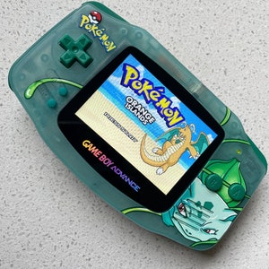 May include: A green Game Boy Advance with a clear shell and a custom design featuring a Pokemon character. The screen displays the game Pokemon Orange Islands with the text "Press Start".