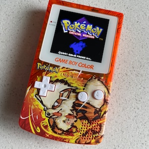 Könnte beinhalten: Orangefarbene Game Boy Color Handheld-Spielkonsole mit einer Pokemon Crystal Version Spielkassette. Die Konsole hat ein individuelles Design mit einem feurigen orangen Hintergrund und einer weißen Pokemon-Figur in der Mitte. Die Konsole hat weiße Knöpfe mit den Beschriftungen A und B, ein Steuerkreuz und einen Startknopf.