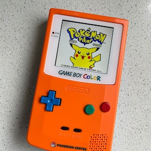 May include: An orange Game Boy Color handheld console displaying the Pokémon Yellow Version game. The console has a blue directional pad, green and red buttons, and the text "GAME BOY COLOR" and "POKEMON CENTER" are visible.