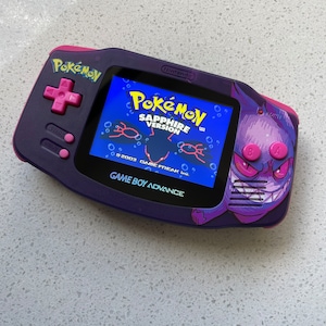 May include: Purple Game Boy Advance with a custom Pokemon Sapphire Version design. The screen shows the game's title card with the text "Pokemon Sapphire Version".