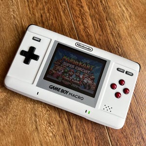 Puede incluir: Consola portátil Game Boy Macro blanca, mostrando el juego Mario Kart: Super Circuit. Cuenta con una cruceta negra, botones rojos y el logotipo de Nintendo. La pantalla muestra un juego de carreras colorido.