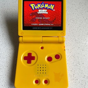 May include: A yellow Game Boy Advance SP handheld gaming console with a red and black Pokemon Ruby Version game cartridge inserted. The console has a red directional pad, red A and B buttons, and a red select and start button.