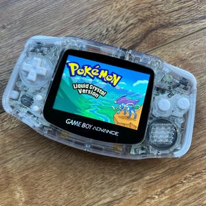 May include: A clear Game Boy Advance handheld gaming console with a translucent white shell. The screen displays the Pokemon Liquid Crystal Version game.