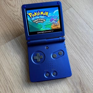 Puede incluir: Consola Game Boy Advance SP azul, abierta para mostrar el juego "Pokémon Liquid Crystal Version". La pantalla muestra un paisaje vibrante con un personaje Pokémon. Los botones y la cruceta de la consola son visibles.