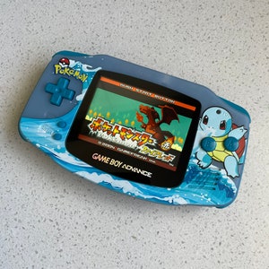 May include: A blue Game Boy Advance handheld console with a Pokémon theme. The console features a screen displaying a Japanese game title. The design includes wave patterns and images of Pokémon characters. The text "GAME BOY ADVANCE" is visible.