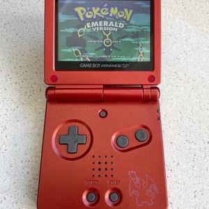 Könnte beinhalten: Eine rote Game Boy Advance SP Handheld-Spielkonsole mit einer Pokemon Emerald Version Spielkassette. Die Konsole hat einen schwarzen Steuerkreuz, zwei schwarze Tasten mit der Aufschrift A und B sowie einen schwarzen Select- und Start-Knopf. Der Bildschirm zeigt den Titelbildschirm von Pokemon Emerald Version.