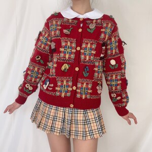 May include: Red knitted cardigan with a plaid pattern and floral embroidery. The cardigan has a button-up front and long sleeves.