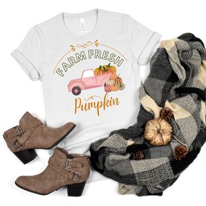 May include: A white t-shirt with the text "FARM FRESH Pumpkin" and a pink truck graphic carrying pumpkins. The shirt is paired with brown ankle boots, and a black and gray plaid blanket with decorative gourds and pinecones.