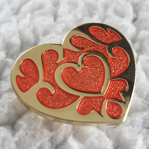 Op de afbeelding: Een gouden hartvormige broche met een ingewikkeld rood en gouden ontwerp. De broche heeft een glanzende afwerking en is perfect om elke outfit een vleugje elegantie te geven.