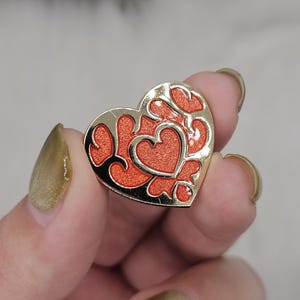 Könnte beinhalten: Eine herzförmige Emaille-Anstecknadel mit einem goldfarbenen Metallrand und filigranen Ausschnitten. Das Innere des Herzens ist mit einer leuchtenden, strukturierten orangefarbenen Glitzer-Emaille gefüllt, die einen Kontrast zum polierten Goldmetall bildet.