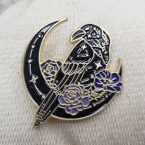 May include: A gold-toned metal enamel pin featuring a black crescent moon and a raven. The raven is decorated with white star patterns, and the design includes purple roses with gold outlines. The pin has a polished finish and intricate details.