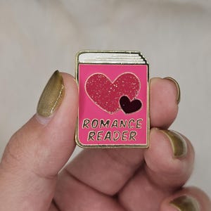 Pode incluir: Um broche de esmalte retangular em forma de livro com capa rosa, apresentando um grande coração vermelho com glitter e um coração escuro menor. O texto ROMANCE READER está impresso em letras douradas, com uma borda de metal dourado.