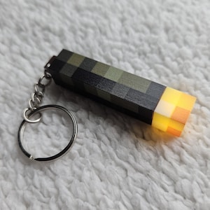 May include: A black and green Minecraft torch keychain with a glowing orange tip. The keychain has a silver metal ring.