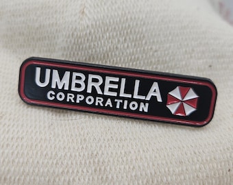 May include: A rectangular black enamel pin featuring the white text UMBRELLA CORPORATION and a red and white umbrella icon on the right side. The pin has a thin red border and a black frame, presenting a clean, high-contrast design for display.