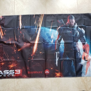 May include: A black and blue flag with a video game character in the center. The character is wearing a black and blue suit with a red and orange arm. The flag has the text "MASS EFFECT 3" in the bottom left corner and "BIOWARE EA" in the bottom right corner.
