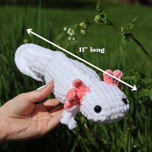 K&ouml;nnte beinhalten: Wei&szlig;es, geh&auml;keltes Axolotl-Pl&uuml;schtier mit rosa Kiemen. Das Spielzeug ist 28 cm lang und wird in einer Hand gehalten.