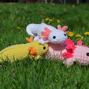 K&ouml;nnte beinhalten: Drei geh&auml;kelte Axolotl-Pl&uuml;schtiere in verschiedenen Farben: Gelb, Wei&szlig; und Rosa. Die Pl&uuml;schtiere liegen auf einem Bett aus gr&uuml;nem Gras.