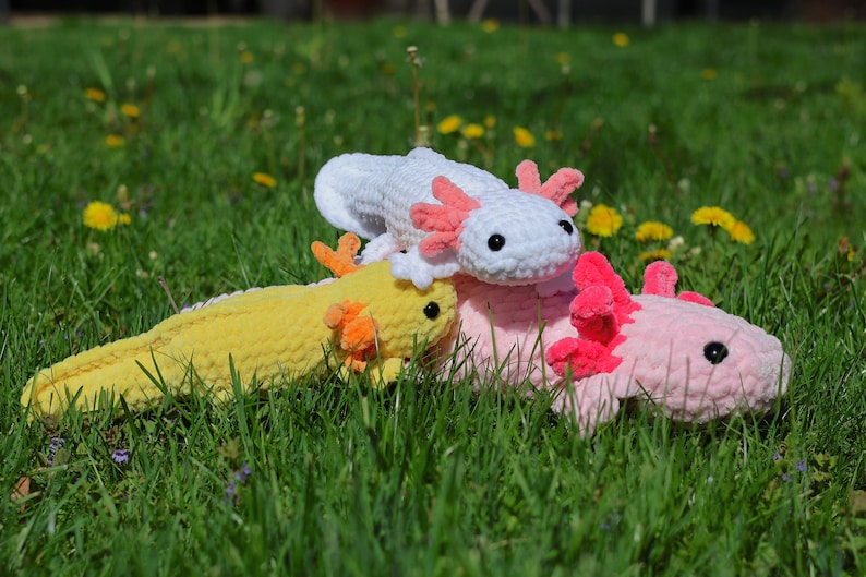 K&ouml;nnte beinhalten: Drei geh&auml;kelte Axolotl-Pl&uuml;schtiere in verschiedenen Farben: Gelb, Wei&szlig; und Rosa. Die Pl&uuml;schtiere liegen auf einem Bett aus gr&uuml;nem Gras.
