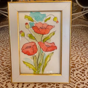 May include: Watercolor painting of red poppies with green leaves and buds, framed in a white frame with a gold inner border. The artwork features a light blue background and is signed in the lower right corner.