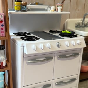 May include: A white toy kitchen stove with black stovetop burners and silver handles. The stove has a shelf with miniature jars and a can of utensils. A small pan with green balls sits on the stovetop. The kitchen is set against a light wood background.