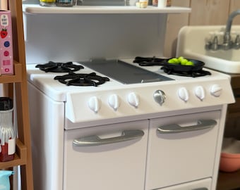 May include: A white toy kitchen stove with black stovetop burners and silver handles. The stove has a shelf with miniature jars and a can of utensils. A small pan with green balls sits on the stovetop. The kitchen is set against a light wood background.