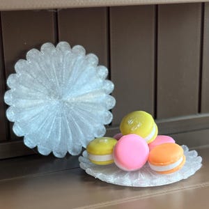 May include: A decorative, silver-colored, scalloped-edge coaster next to a plate of colorful macarons. The macarons are yellow, pink, and orange with white filling. The background is a brown, vertical-paneled surface.