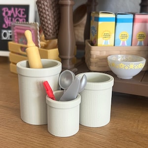 May include: A selection of miniature kitchen items, including three off-white ceramic pots, a rolling pin, and utensils. The pots vary in size and texture, with one having vertical ridges. A small bowl with yellow floral designs is also present.
