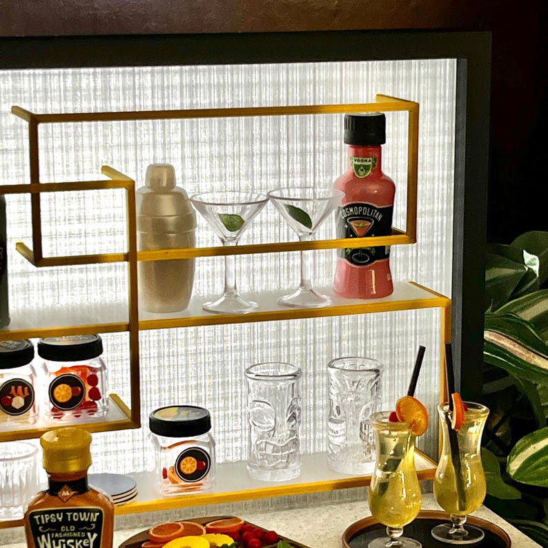 Op de afbeelding: Een miniatuur barset met een gouden frame en planken. De planken zijn gevuld met miniatuur flessen drank, cocktailshakers, glazen en potten met fruit. De barset is van achteren verlicht met wit licht.
