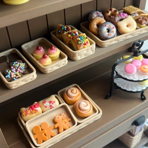 Puede incluir: Una exhibición de panadería en miniatura con varios productos horneados. La exhibición incluye cupcakes, donas, rollos de canela y galletas, todos dispuestos en pequeñas cestas tejidas y en un soporte escalonado. Los pasteles están decorados con glaseado y chispas de colores.