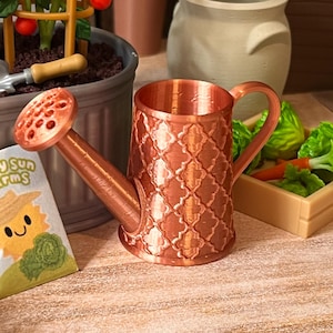 May include: A miniature copper-colored watering can with a decorative pattern. The can has a long spout and a round sprinkler head. The image also shows a small garden scene with a tomato plant, a small trowel, and a container of vegetables.