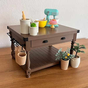 Puede incluir: Una isla de cocina marrón en miniatura con un cajón y un estante inferior, que presenta una variedad de pequeños accesorios de cocina. Incluye una batidora turquesa, un cuenco amarillo, un pastel rosa y pequeñas plantas en macetas. Una pequeña cesta cuelga a un lado.