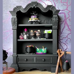 May include: A black wooden hutch with two drawers and three shelves. The shelves are decorated with Halloween themed items, including a cauldron, sweets, and desserts. The hutch is against a purple and pink background with a tree branch design.