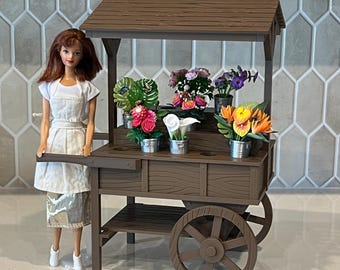 May include: A doll with auburn hair wearing a white apron and skirt stands beside a brown plastic flower cart. The cart features a roof, two wheels, and multiple shelves holding miniature potted plants, including pink, white, and orange flowers.