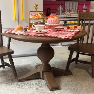 May include: A miniature dining scene with a round wooden table, set with a red and white checked tablecloth. The table is laden with miniature desserts, a tea set, and a tiered serving tray. Two wooden chairs flank the table, creating a cosy setting.