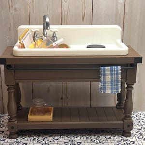 May include: A miniature kitchen sink with a white basin and chrome tap sits on a brown wooden cabinet. The sink is filled with toy dishes. A blue and white checked tea towel hangs below. A wooden tray and glass jar are on the lower shelf.