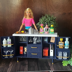 Op de afbeelding: Een blauwe en gouden barwagen met planken en lades. De wagen is gevuld met miniatuurflesjes drank, glazen en bargereedschap. Een pop met een roze shirt staat achter de wagen.
