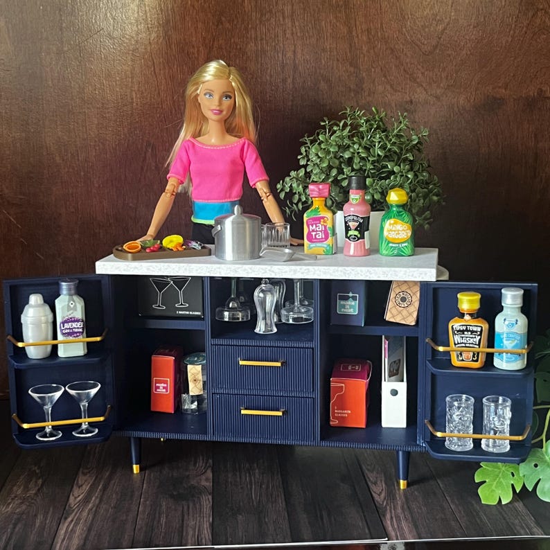 Op de afbeelding: Een blauwe en gouden barwagen met planken en lades. De wagen is gevuld met miniatuurflesjes drank, glazen en bargereedschap. Een pop met een roze shirt staat achter de wagen.