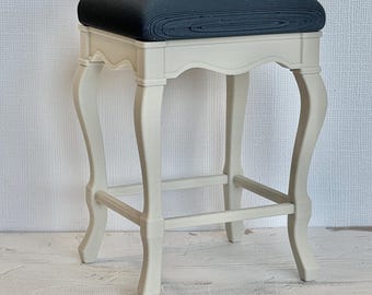 May include: A small stool featuring a rectangular black upholstered seat cushion atop a cream-colored frame. The base consists of four curved legs connected by thin horizontal support bars, creating a stable structure for this piece of furniture.