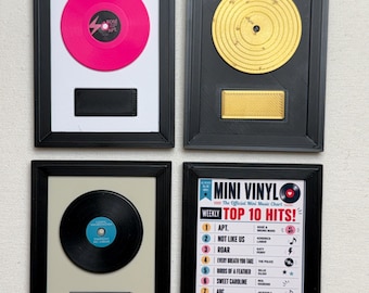 May include: Four framed wall art pieces featuring miniature vinyl records. Three frames contain a single record in hot pink, gold, or black with a plaque below. The fourth frame displays a printed list titled Mini Vinyl Weekly Top 10 Hits.