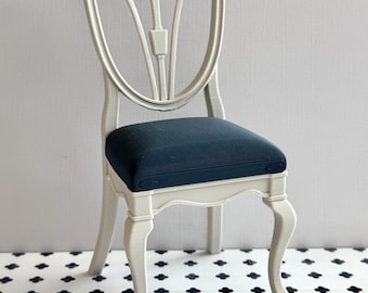 May include: A miniature chair featuring a light cream-colored frame with a shield-shaped backrest and curved legs. The seat is upholstered in a dark navy blue fabric, providing a contrast to the light-colored structure of the piece.