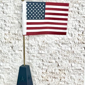 May include: A miniature American flag with red and white stripes and a blue field of white stars. The flag is mounted on a gold-colored pole and set in a dark blue, star-adorned base. The background is a white brick wall.