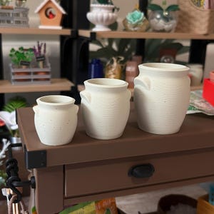 May include: Three off-white ceramic pots of different sizes. Each pot features a textured finish and two small handles. The pots are displayed on a brown surface, possibly for decorative purposes. The pots are likely for plants or other decorative items.