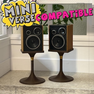 May include: Two miniature speakers with black fronts and brown sides, each mounted on a dark brown pedestal stand with a gold accent ring. A yellow speech bubble graphic at the top contains the text MGA's MINI VERSE COMPATIBLE.