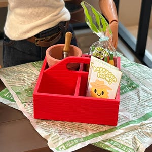 May include: A red wooden caddy with compartments, holding a small mortar and pestle, a bag of miniature carrots, and a card that says "Happy Sun Farms" with a smiling sun graphic. The caddy sits on a newspaper.