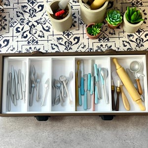 Könnte beinhalten: Eine braune Holzschublade gefüllt mit verschiedenen Miniatur-Küchenutensilien, darunter Messer, Gabeln, Löffel und ein Nudelholz. Die Schublade ist in Fächer unterteilt, und die Utensilien sind ordentlich angeordnet. Kleine Topfpflanzen und Behälter sind im Hintergrund zu sehen.