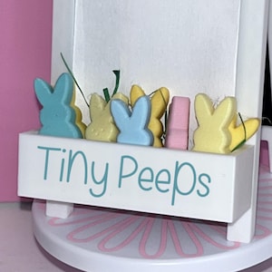 Könnte beinhalten: Eine weiße Holzkiste mit den Worten "Tiny PeepS" in blaugrüner Farbe. Die Kiste ist gefüllt mit pastellfarbenen, kaninchenförmigen Marshmallow-Süßigkeiten.