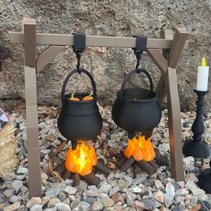 Puede incluir: Calderos negros en miniatura colgados de un marco de madera marrón, con llamas y troncos naranjas debajo. Un caldero contiene objetos naranjas. Un candelabro negro con una vela blanca está a la derecha. La escena está sobre un lecho de pequeñas rocas.
