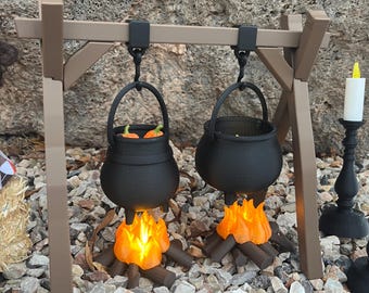May include: Miniature black cauldrons hanging from a brown wooden frame, with orange flames and logs beneath. One cauldron contains orange objects. A black candlestick with a white candle is to the right. The scene is set on a bed of small rocks.